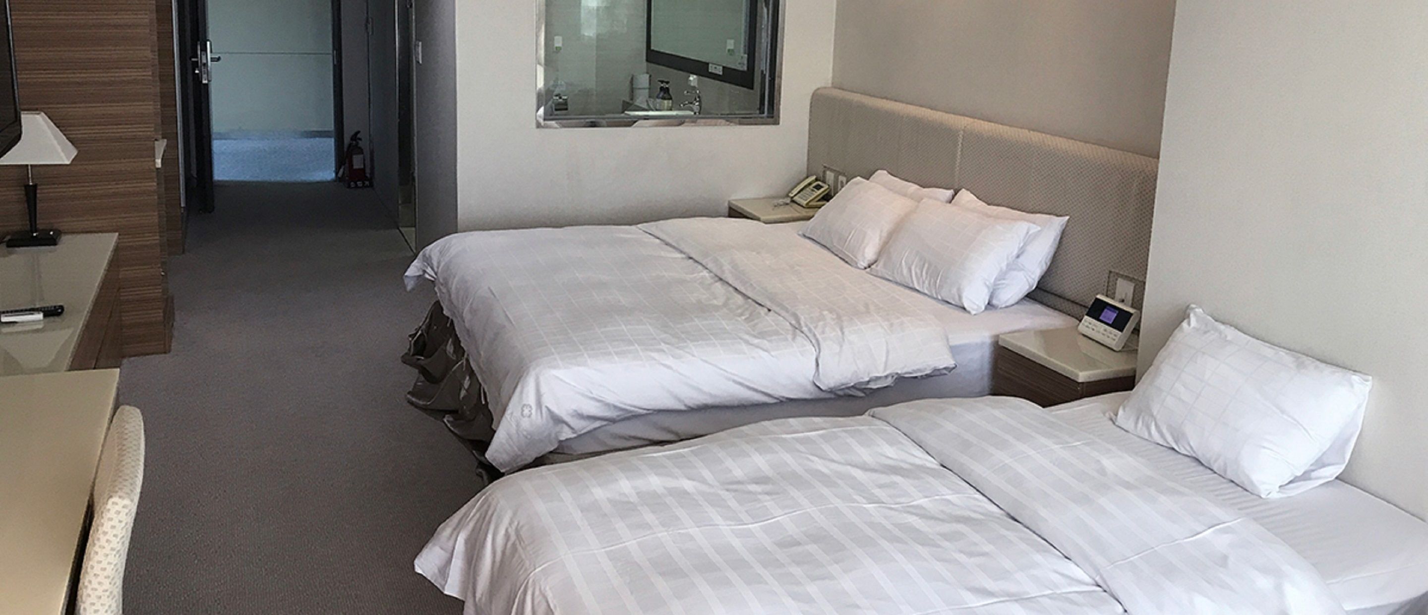 A hotel room with two beds, a bedside table with a lamp, a mirror, and a small bedside table with a phone and a digital device.