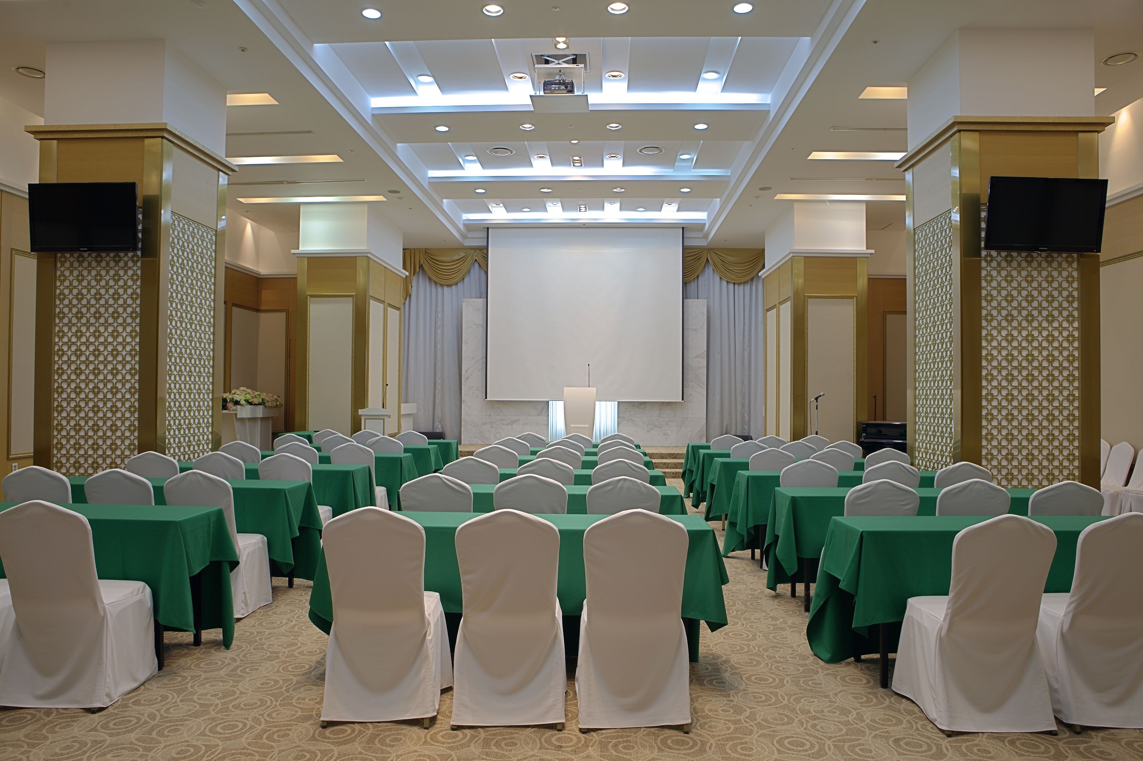 Un salón de conferencias con filas de mesas y sillas, una pantalla grande y un atril.