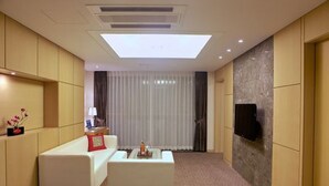 A hotel room with a sofa, a coffee table, a flat-screen TV mounted on the wall, and a large window with curtains. - The Class 300 (Sokcho)