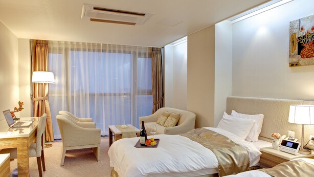 A hotel room with a bed, two chairs, a desk with a laptop, a bedside table with a lamp, and a window with curtains. - The Class 300 (Sokcho)