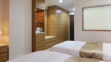 A hotel room with two beds, a wooden wardrobe, a bedside table with a lamp, and a window with blinds.
