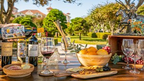 Mesa posta com garrafas de vinho, copos, uma tigela de madeira com frutas e uma vista panorâmica ao ar livre.