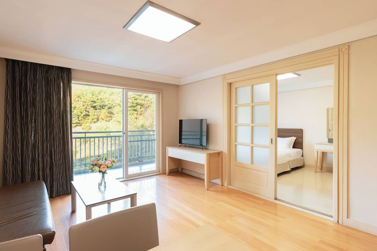 Une chambre d’hôtel moderne avec un plancher en bois, un bureau doté d’un téléviseur à écran plat, un lit, une porte coulissante et un balcon offrant une vue sur la verdure.