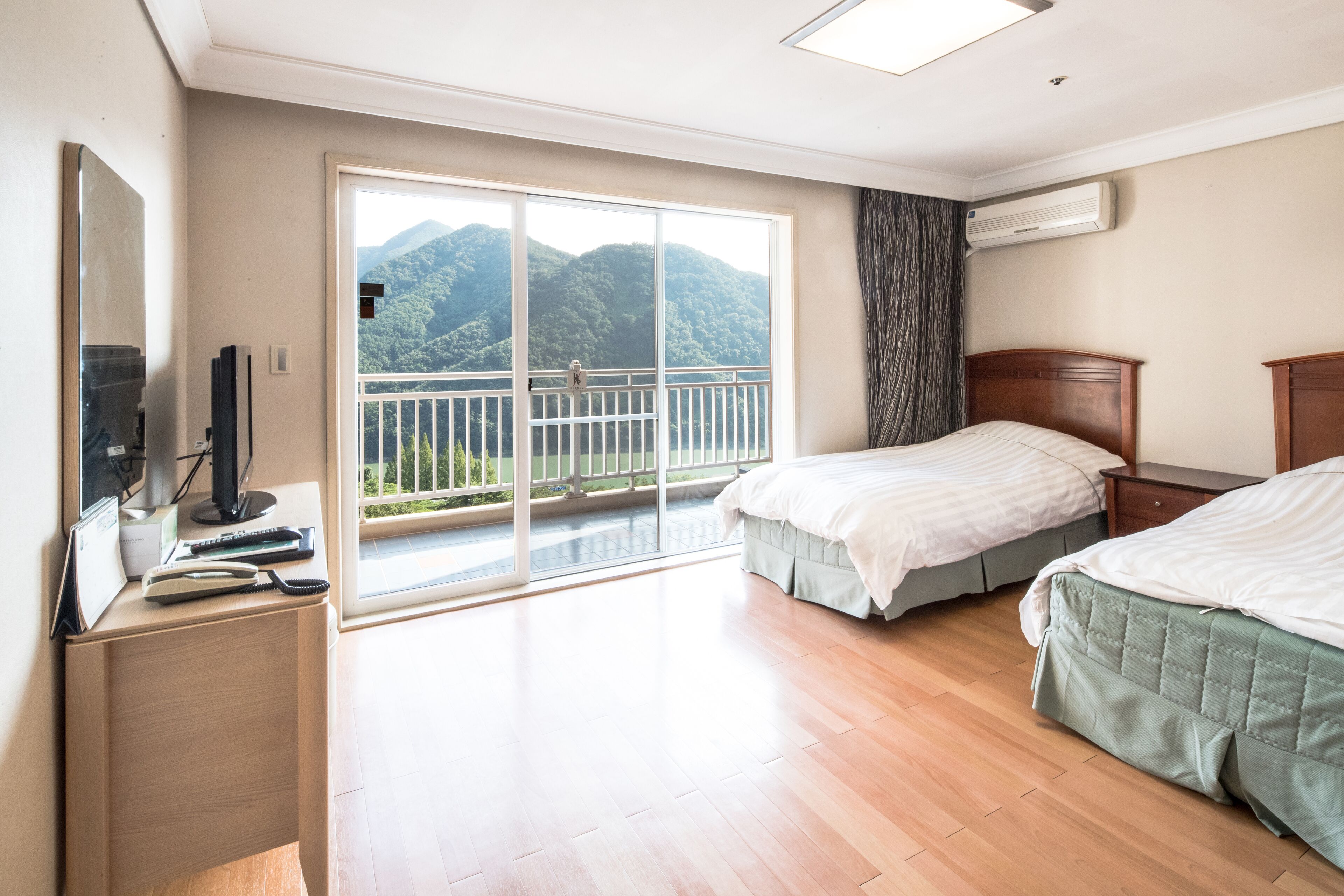 A bedroom with two beds, a desk with a computer, and a large sliding glass door leading to a balcony with a mountain view.