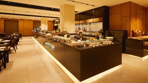 Uma área de buffet com várias estações de comida, uma cozinha ao fundo e diferentes opções de assentos.