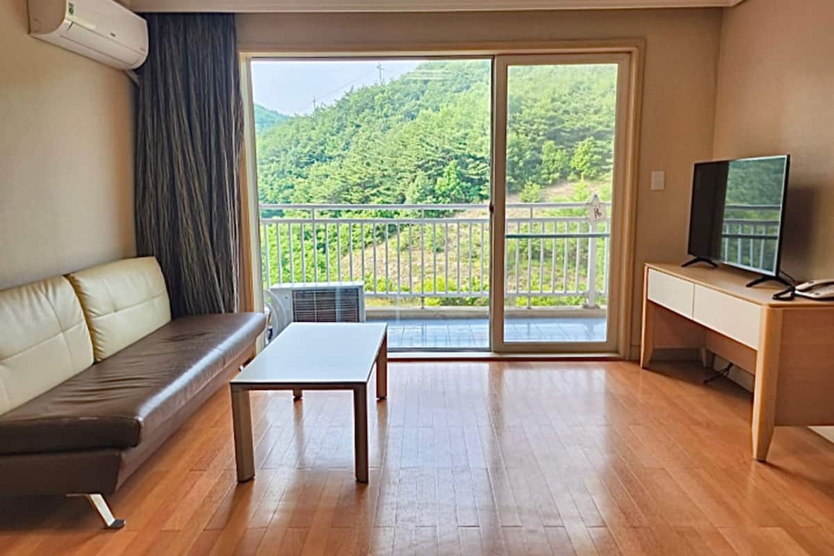 Une pièce comprenant un canapé, une petite table, un bureau avec une télévision et un balcon donnant sur un paysage verdoyant.