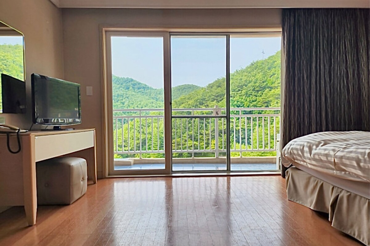 A hotel room with a wooden floor, a bed, a desk with a television, and a balcony with a view of green hills.