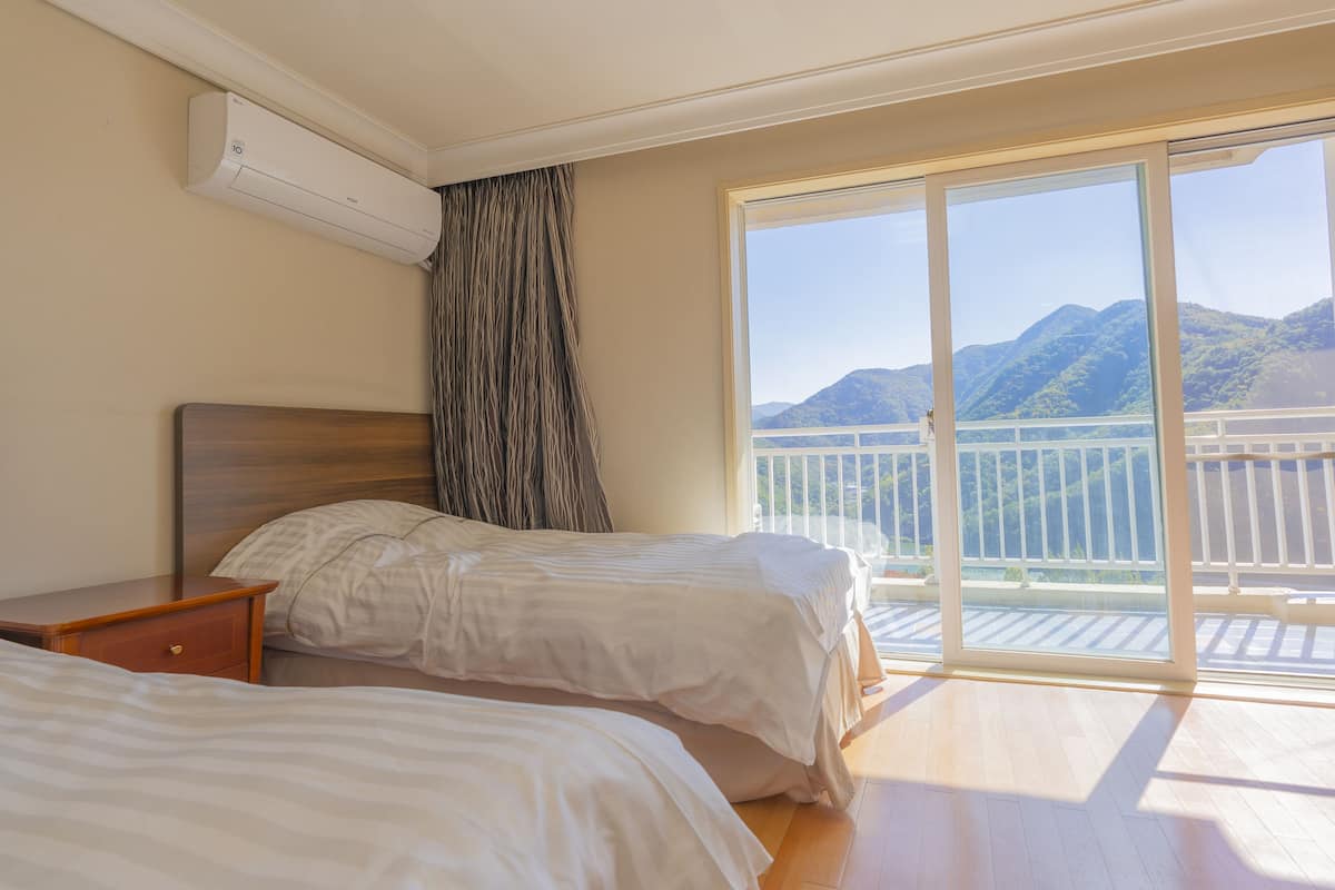 Une chambre d’hôtel avec deux lits, une tête de lit en bois, une table de chevet et une grande porte-fenêtre coulissante donnant sur un balcon avec vue sur la montagne.