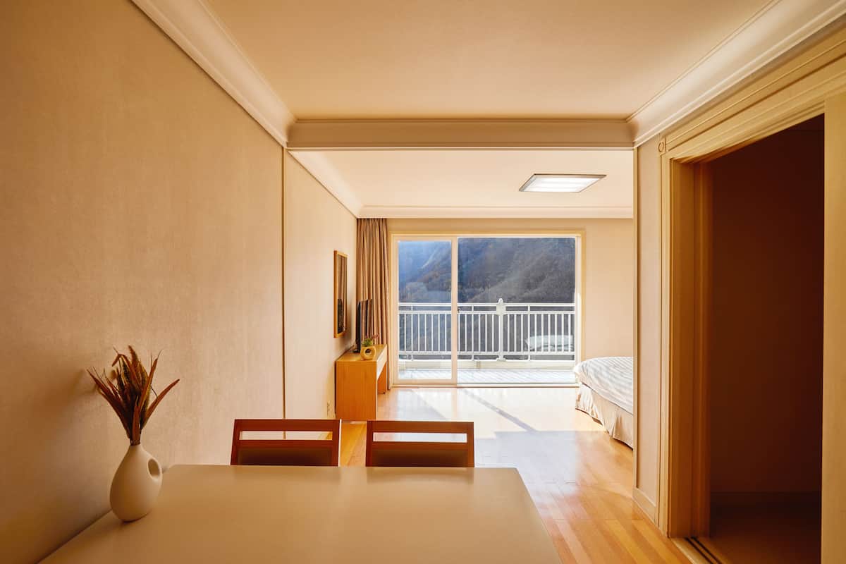 Un coin repas avec une table, des chaises et un vase contenant des plantes séchées. Il y a un balcon avec un lit et une vue sur l’extérieur.
