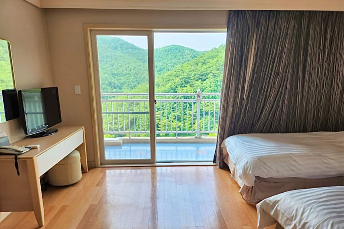 Une chambre d’hôtel avec un plancher en bois, un lit, un bureau avec une télévision et un balcon offrant une vue sur des collines verdoyantes.