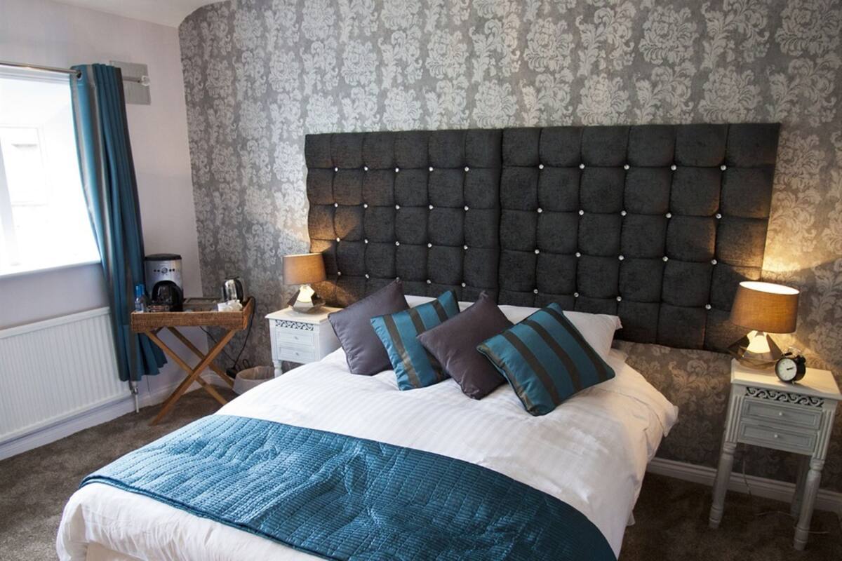A bedroom with a tufted headboard, a bed with a blue and white bedspread, two bedside tables with lamps, a small desk with a lamp, and a window with curtains.