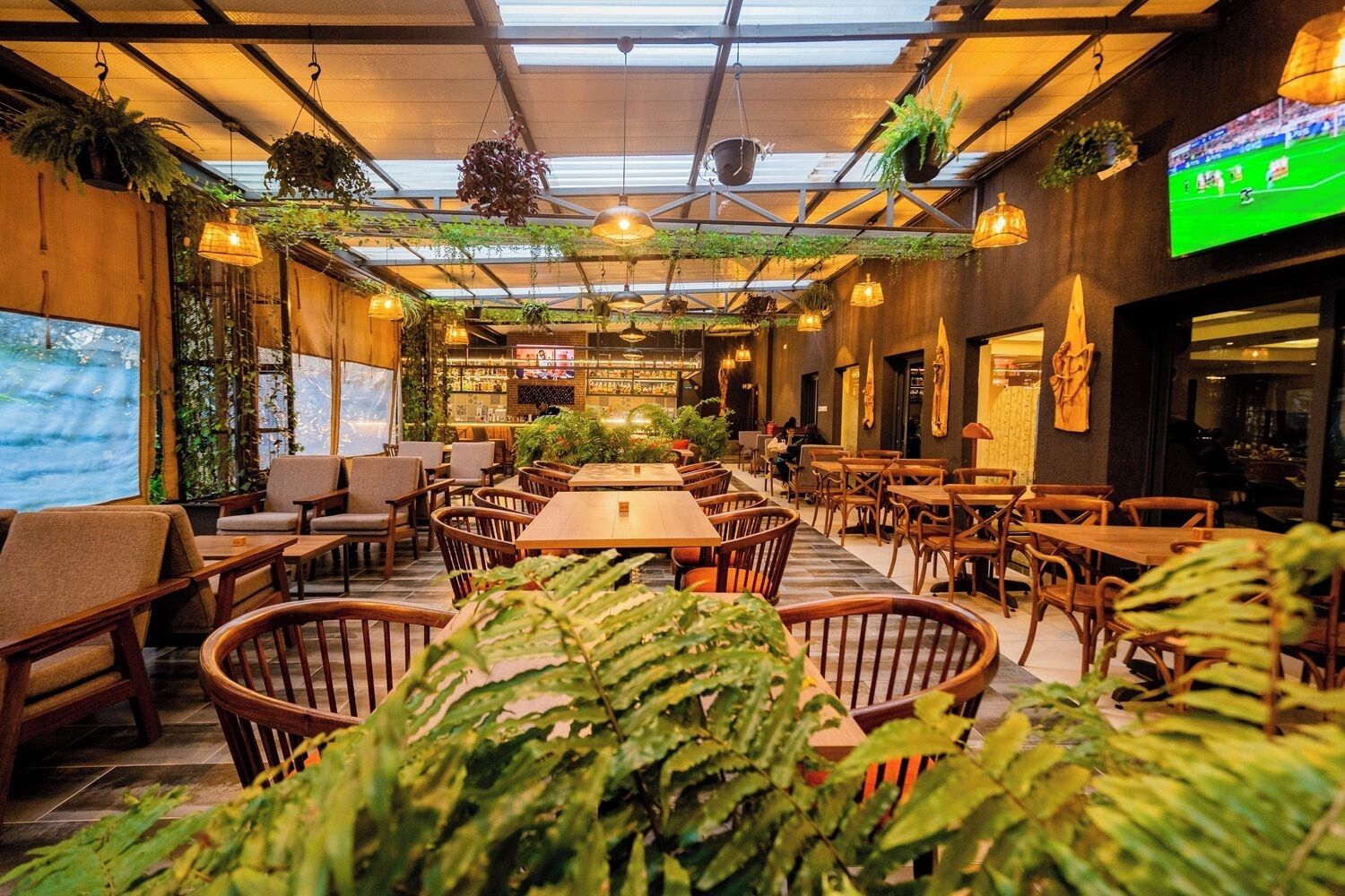 An indoor restaurant with wooden tables and chairs, hanging plants, and a television screen.