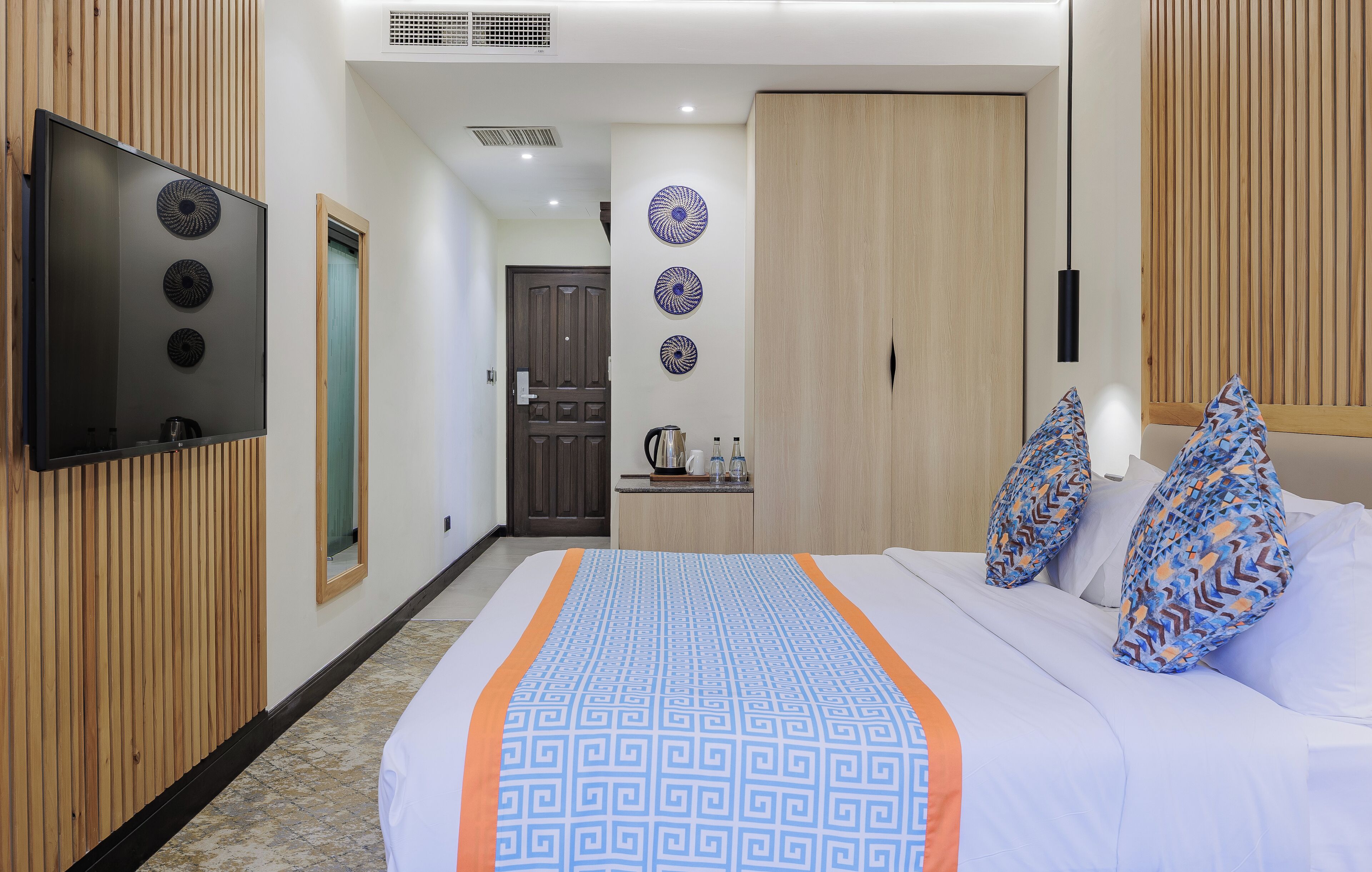 A hotel room with a bed, a TV mounted on the wall, a wooden wardrobe, and a small table with a kettle and cups.