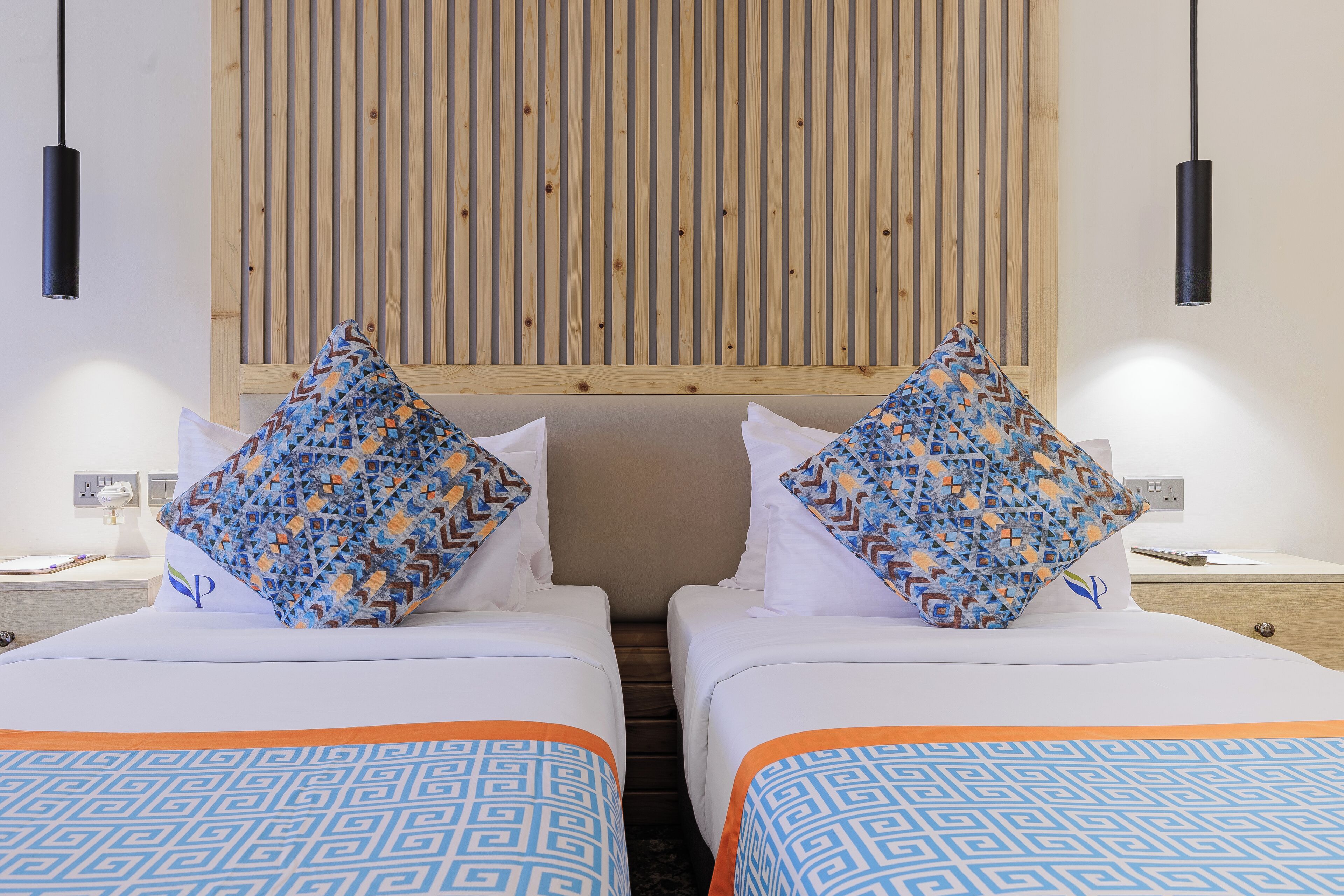 A hotel room with two beds, patterned pillows, a wooden slat headboard, and pendant lights.