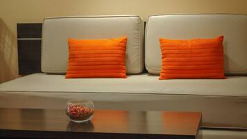 Una habitación con un sofá, dos almohadas naranjas, un cuadro en la pared y una mesa de centro con un bol de caramelos.