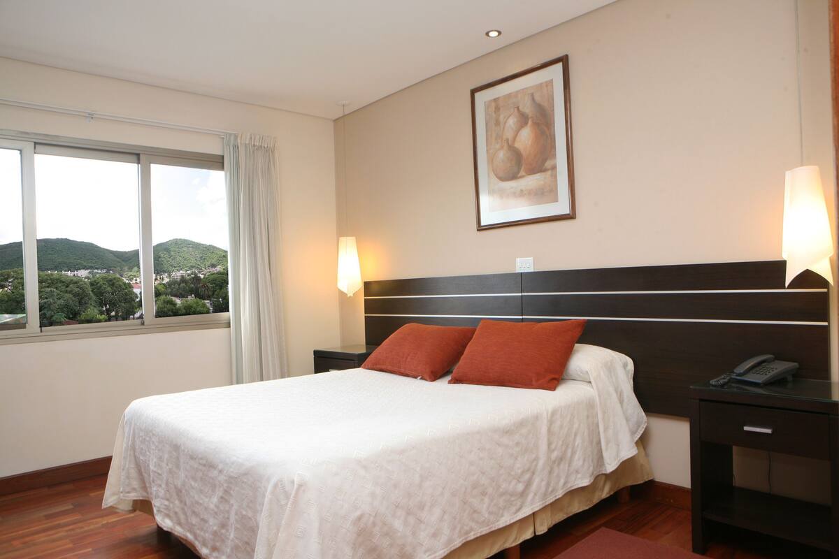 A hotel room with a large bed, two bedside lamps, a nightstand with a phone, and a view of greenery through the window.