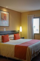 Habitación de hotel con cama, mesita de noche, lámpara, ventana con cortinas y un cuadro en la pared.