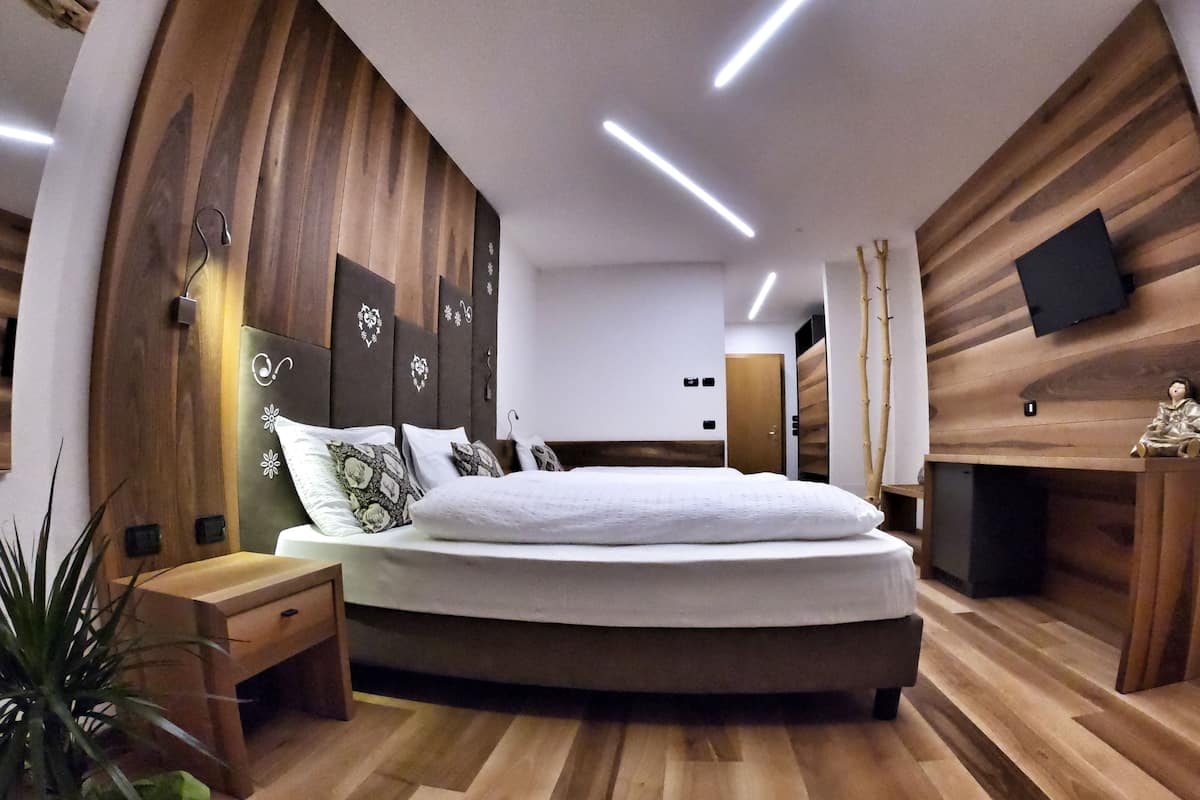 Un dormitorio moderno con cabecera de madera, una cama grande con muchos almohadones, una mesita de noche y un televisor montado en la pared.
