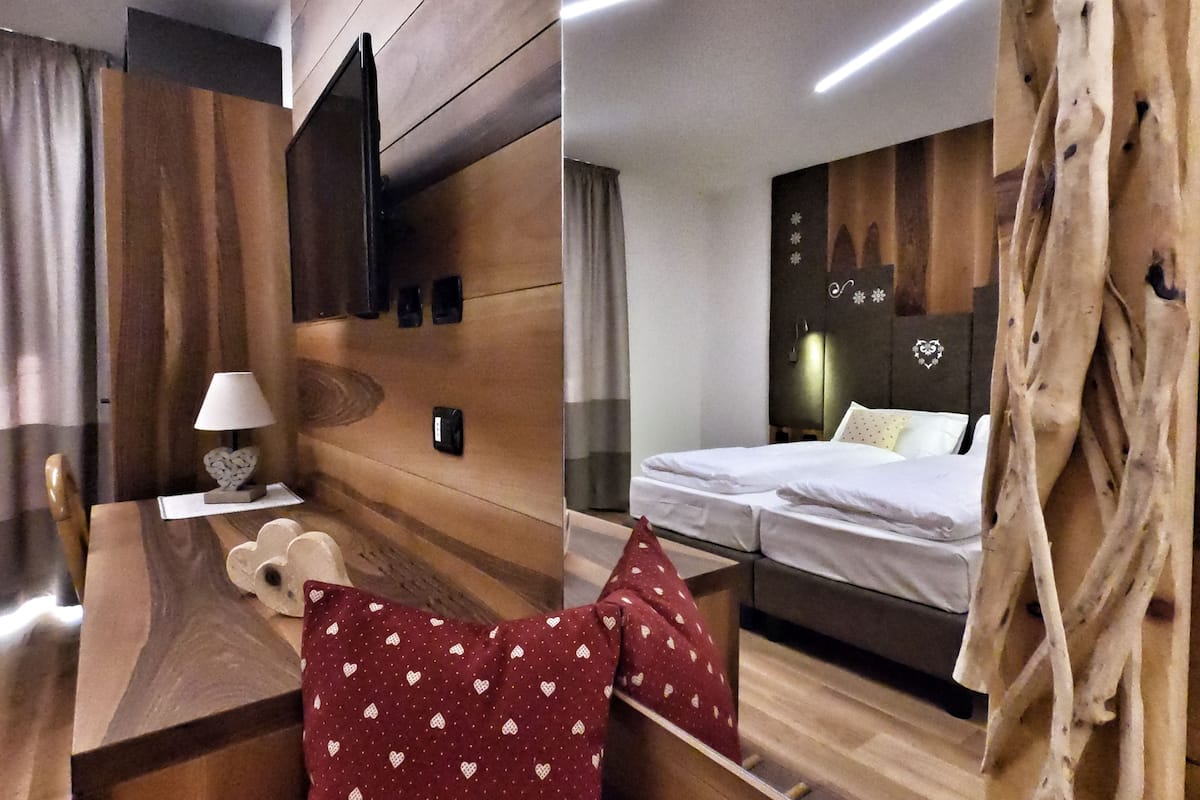Habitación de hotel con pared de madera, cama con ropa de cama blanca, escritorio con lámpara y almohada con diseño de corazones rojos.