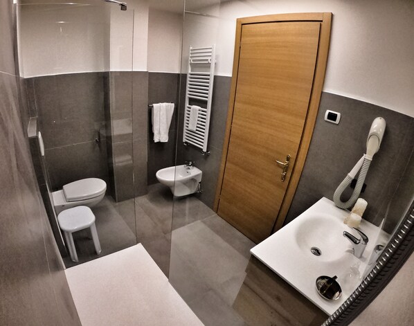 A modern bathroom with a glass-enclosed shower, a bidet, a toilet, a sink with a mirror, and a wooden door. - Hotel Cristallo (Canazei)