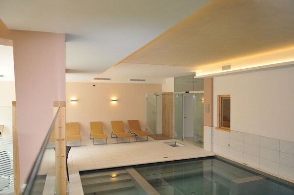Un área moderna de piscina interior con pared de vidrio, zona de estar con sillas amarillas y un pilar de madera.