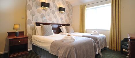 A hotel room with two beds, a nightstand with a lamp, a dresser, and a window with curtains.