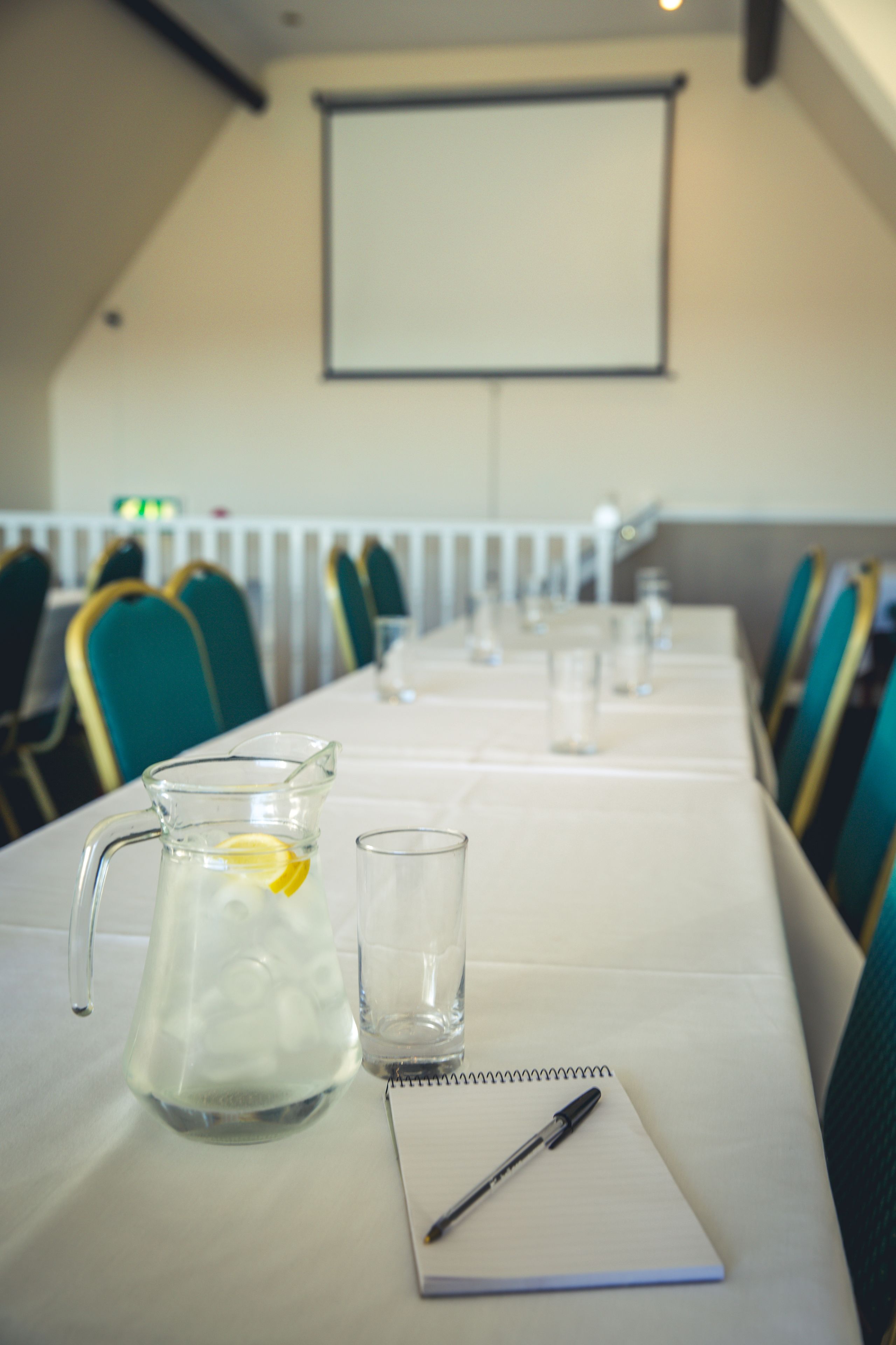 A conference room with a long table, chairs, a projector screen, a pitcher, a glass, and a notepad.