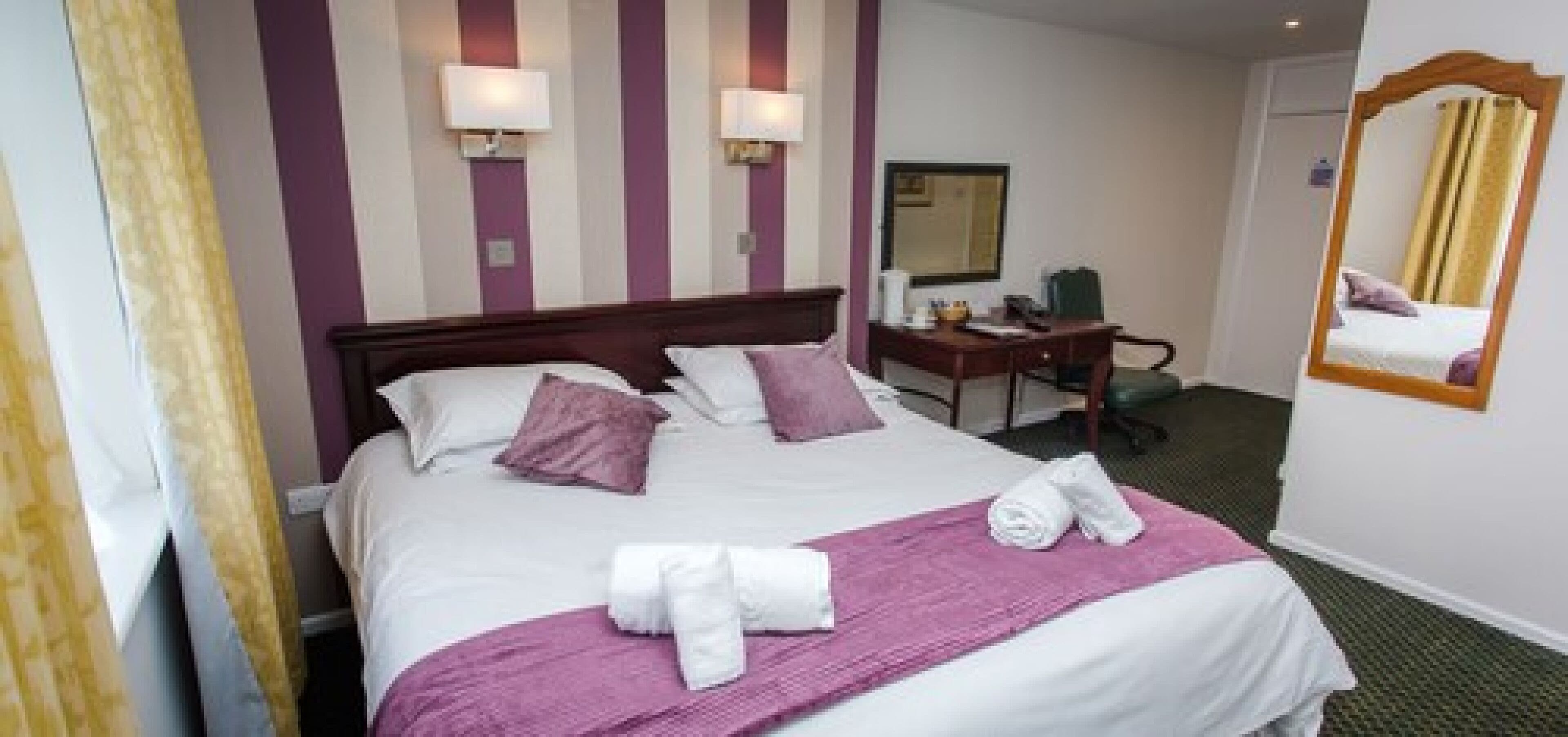 A hotel room with a large bed, two bedside lamps, a desk with a chair, a mirror, and a window with curtains.