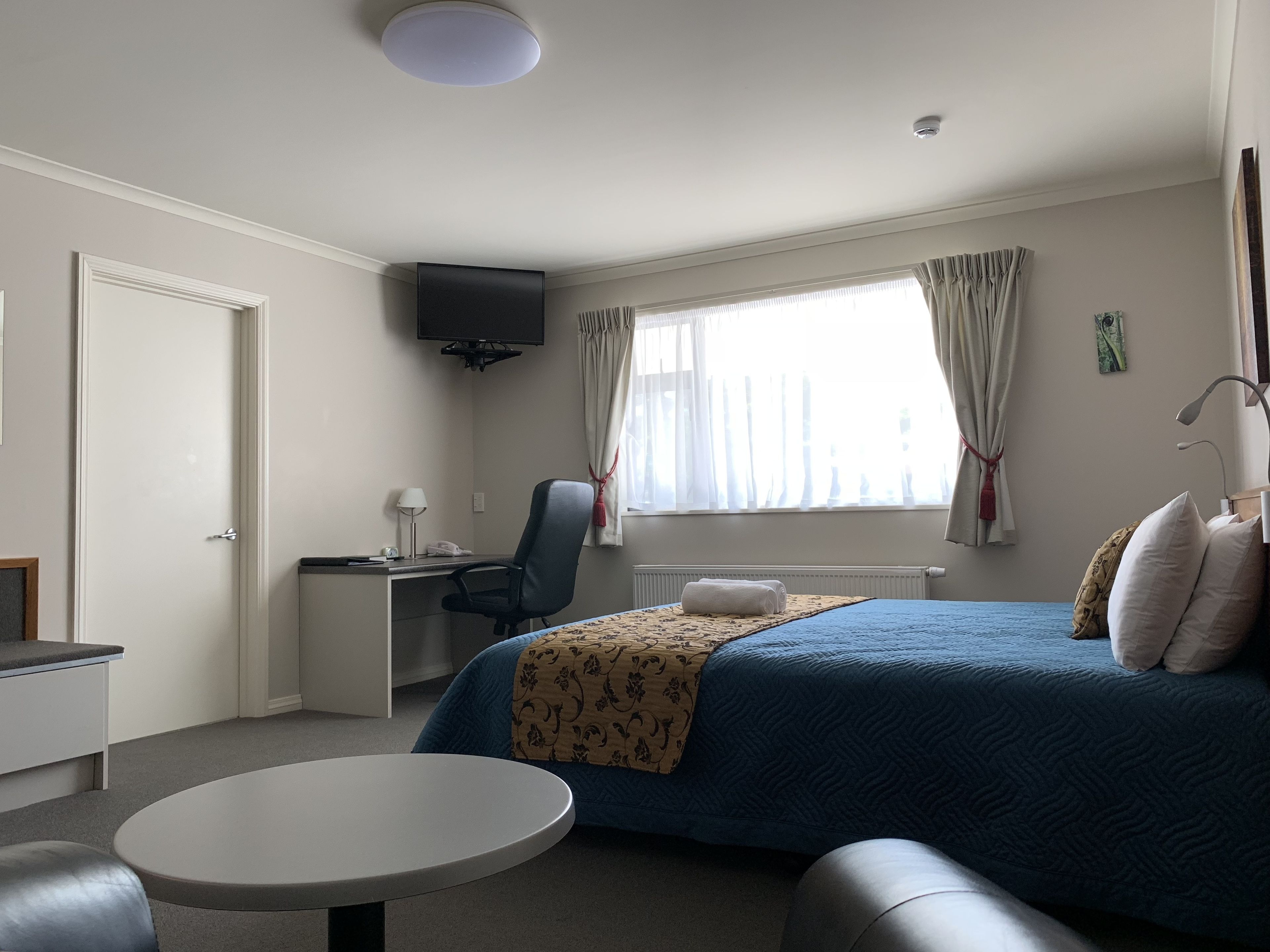 A hotel room with a bed, a desk, a chair, a small table, and a TV on the wall.