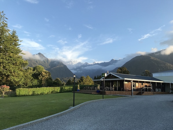 A building with a gravel driveway and a lawn, surrounded by trees and mountains in the background. - High Peaks Hotel (Fox Glacier)