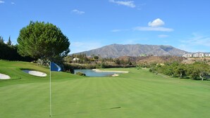 Un campo de golf con un mástil de bandera, búnkeres de arena, un estanque y una montaña al fondo.