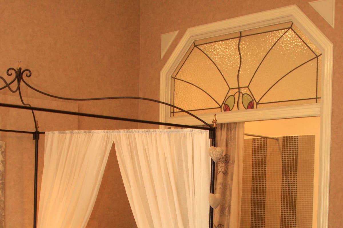 A four-poster bed with a canopy, a bedside table with a lamp, a mannequin with a heart design, and a bathroom with a shower and a small stool.