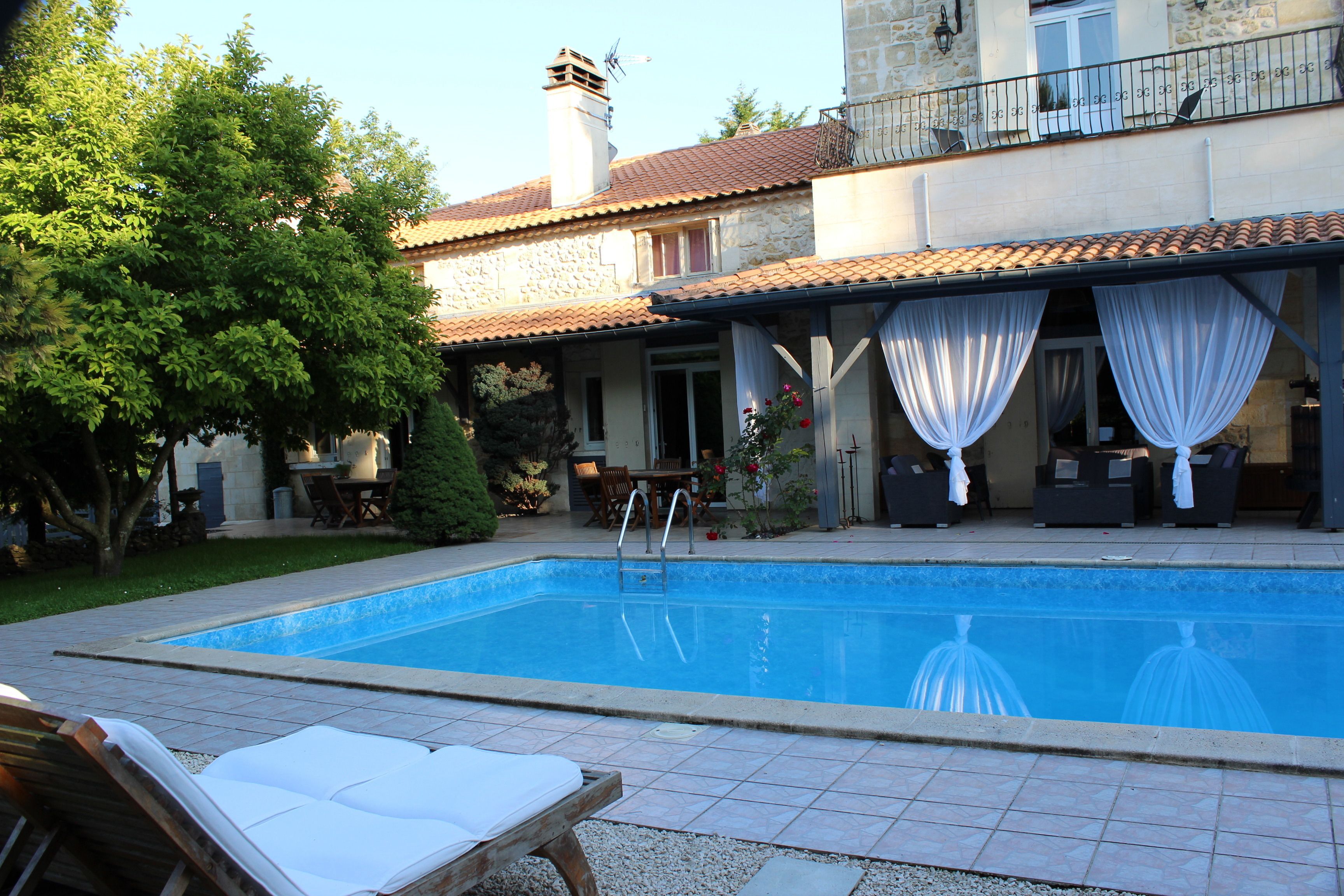 A swimming pool with a tiled deck, lounge chairs, and a covered patio area with white curtains.