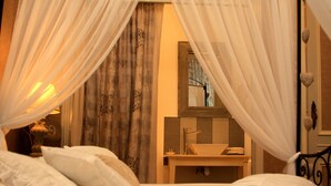 Un dormitorio con cama, almohadas, mesita de noche, espejo, lavamanos y ventana con cortinas.