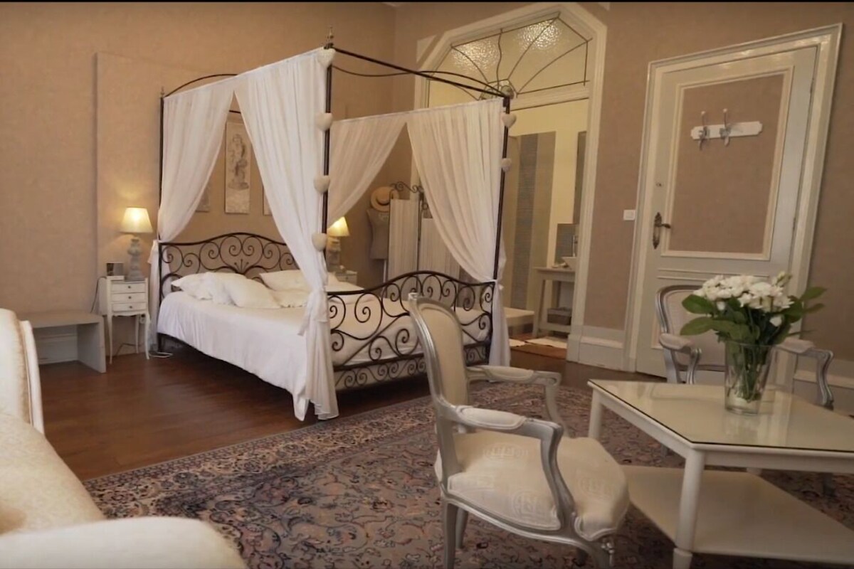 Un lit à baldaquin à quatre poteaux, une table de chevet, un fauteuil confortable, une petite table avec un vase de fleurs et une chaise.