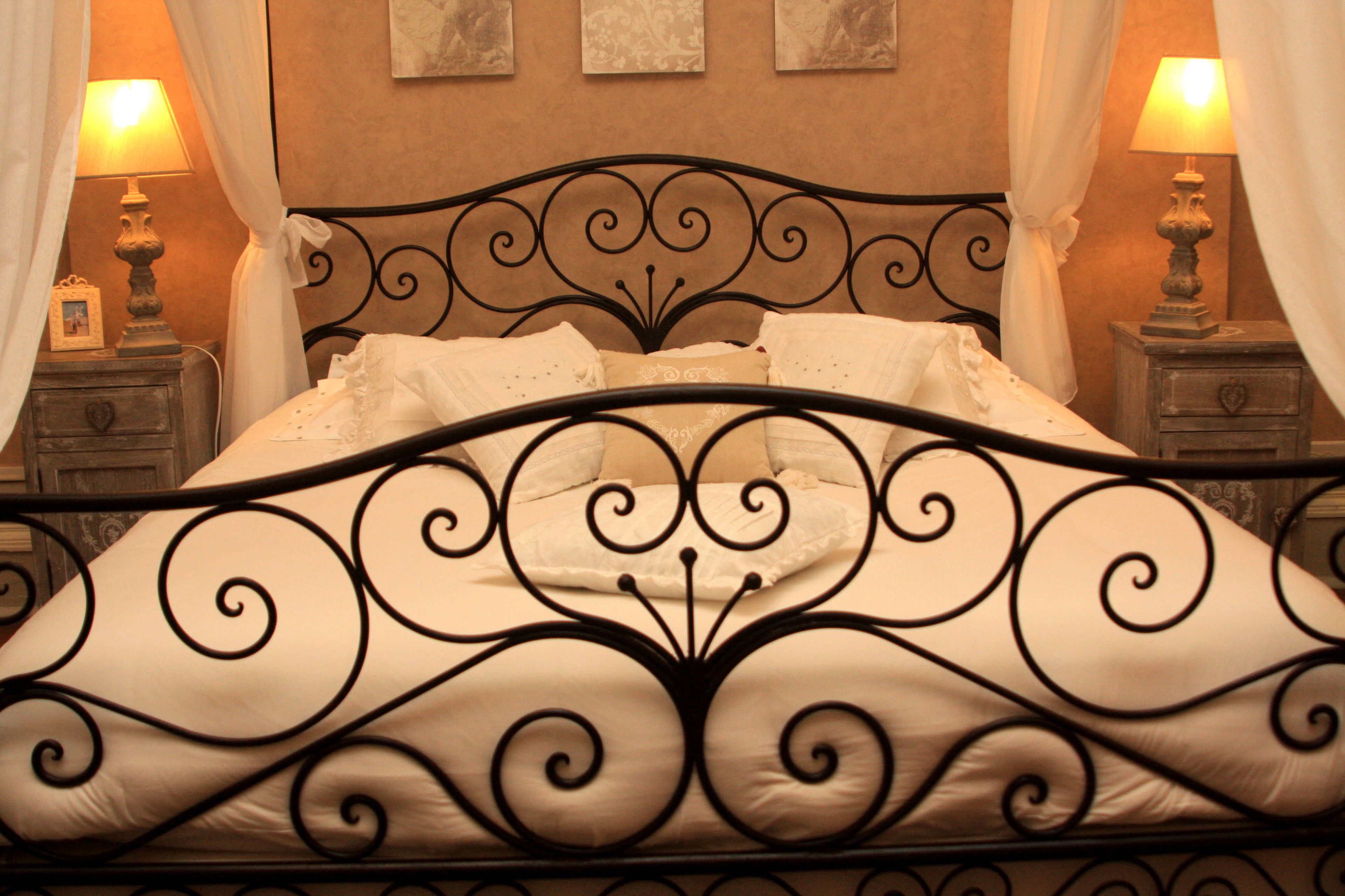 Una cama con estructura metálica negra y ropa de cama blanca, flanqueada por dos mesitas de noche con lámparas y cuadros enmarcados.