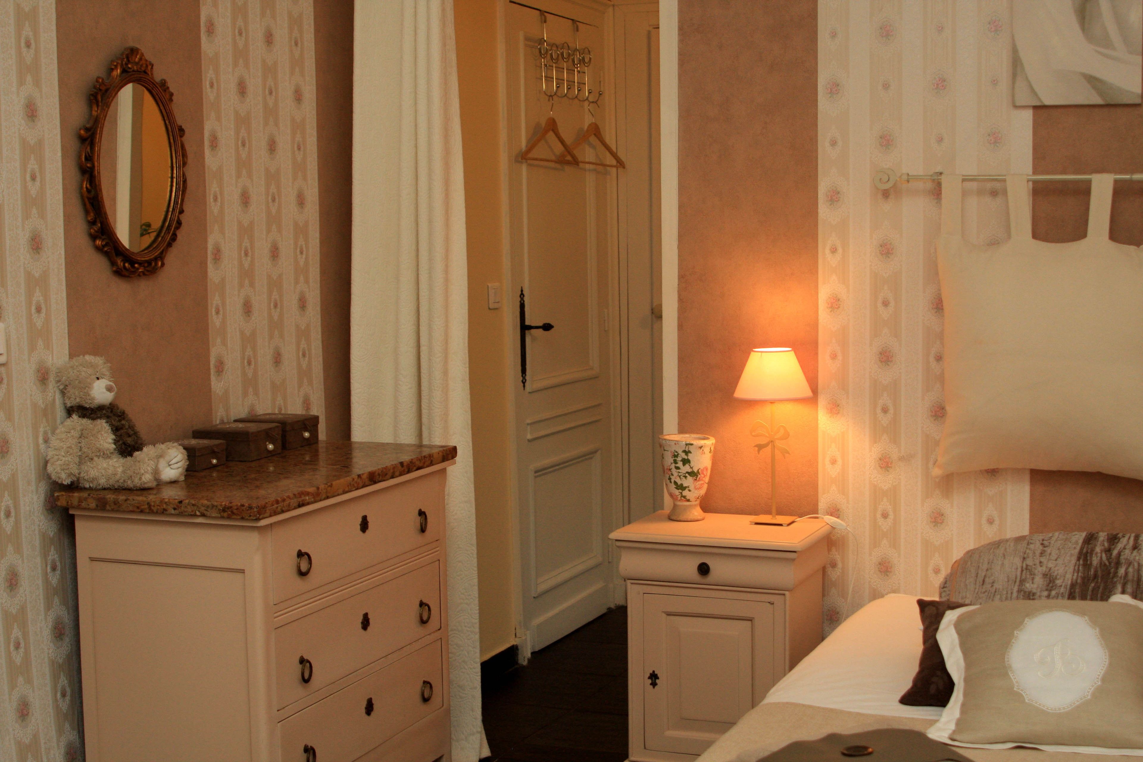 A bedroom with a dresser, a bedside table with a lamp, a bed with a pillow, a mirror, and a hanging coat rack.