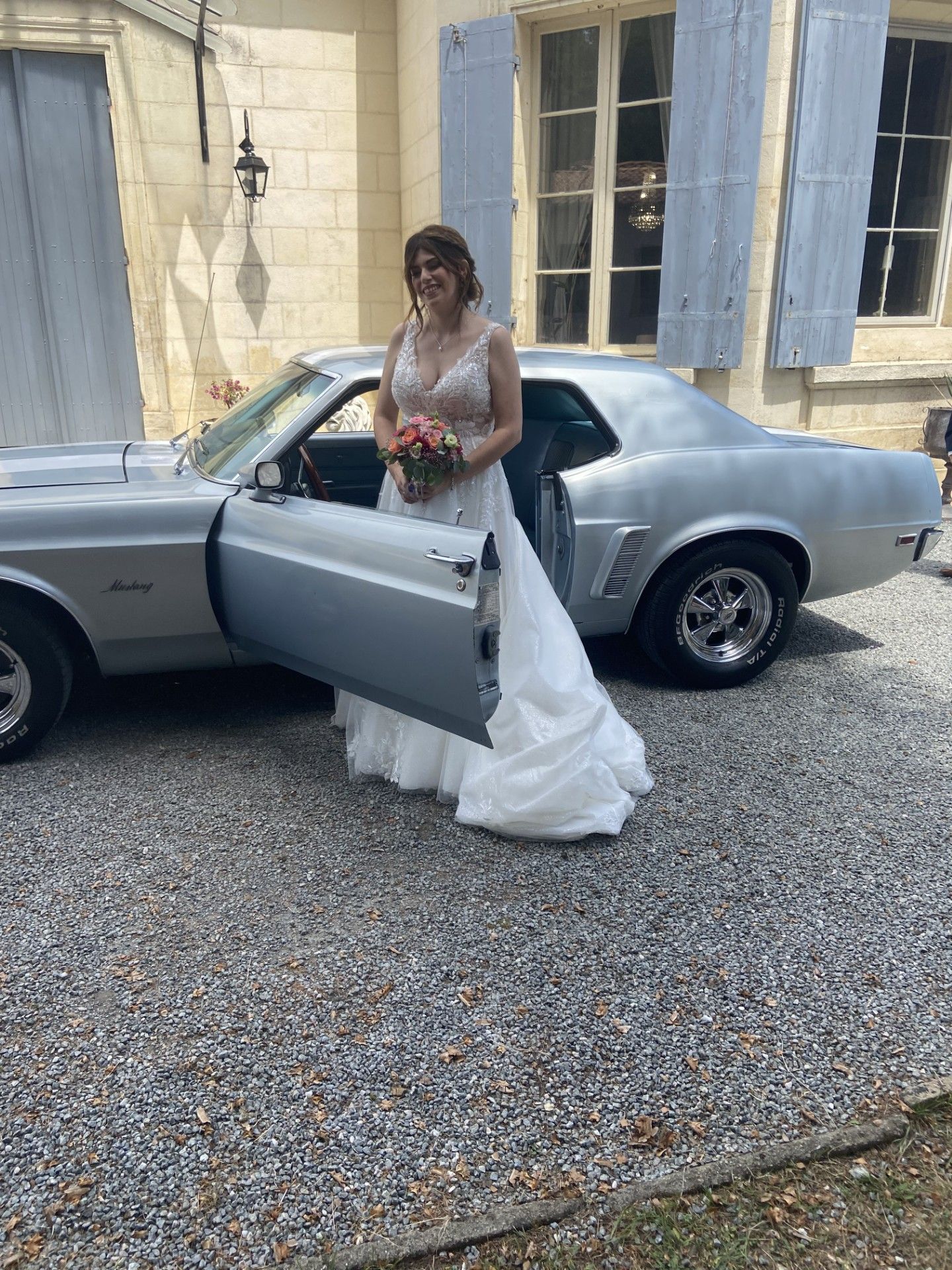 Escena de boda con una novia en vestido blanco posando junto a un coche antiguo.
