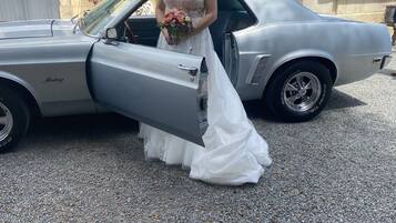 Escena de boda con una novia en vestido blanco posando junto a un coche antiguo.