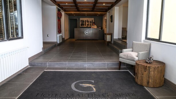 Uma área de recepção de hotel com balcão, sala de espera com uma cadeira e mesa redonda de madeira, e tapete preto com o logotipo do hotel.