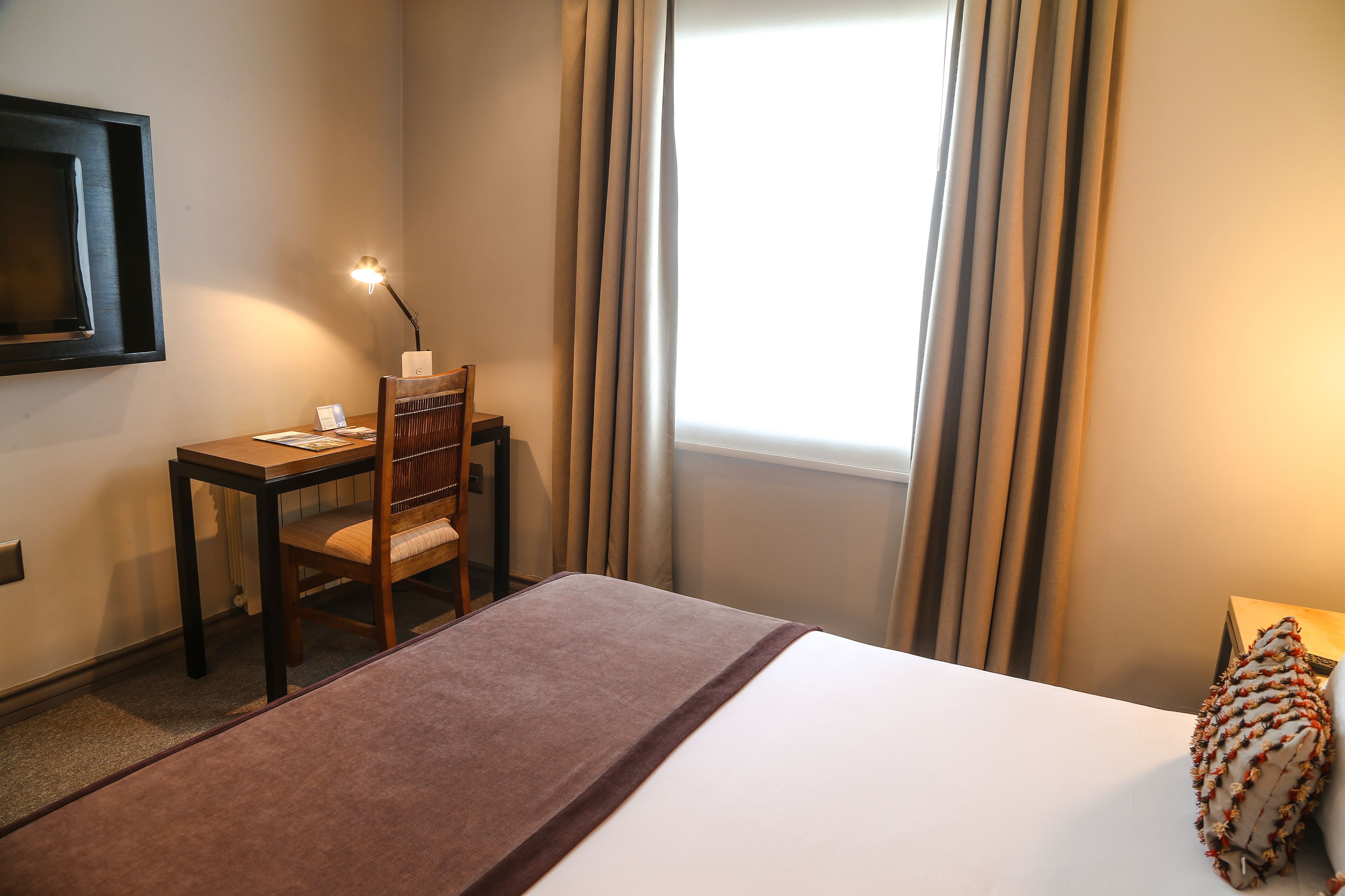 Habitación de hotel con cama, un escritorio con silla, una lámpara y una ventana con cortinas.