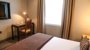 Habitación de hotel con cama, un escritorio con silla, una lámpara y una ventana con cortinas.