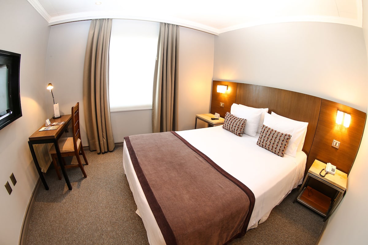 Une chambre d’hôtel avec un grand lit, un bureau avec une chaise, une table de chevet avec une lampe et une vue sur l’extérieur par une fenêtre munie de rideaux.