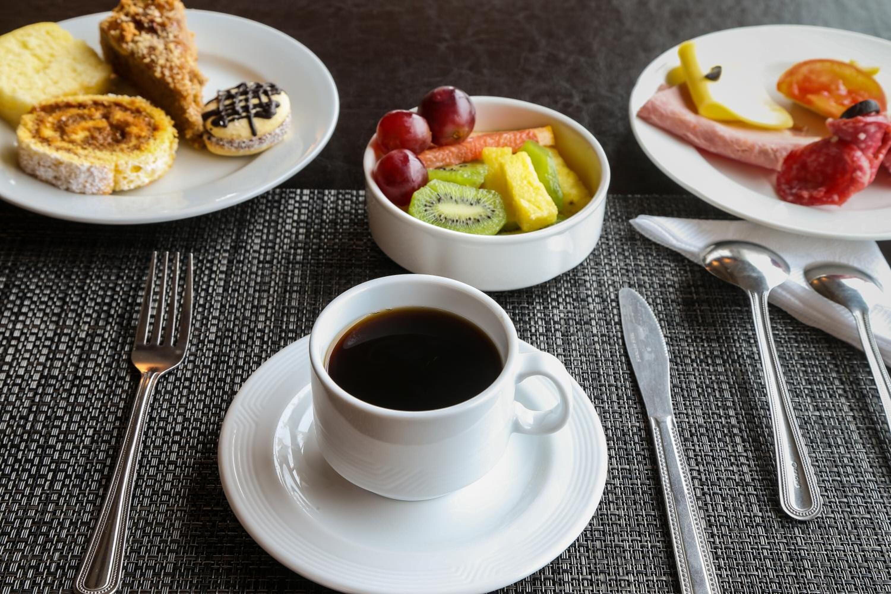 Une table mise pour le petit-déjeuner avec une tasse de café, des assiettes de toasts, des fruits et une tranche de jambon.