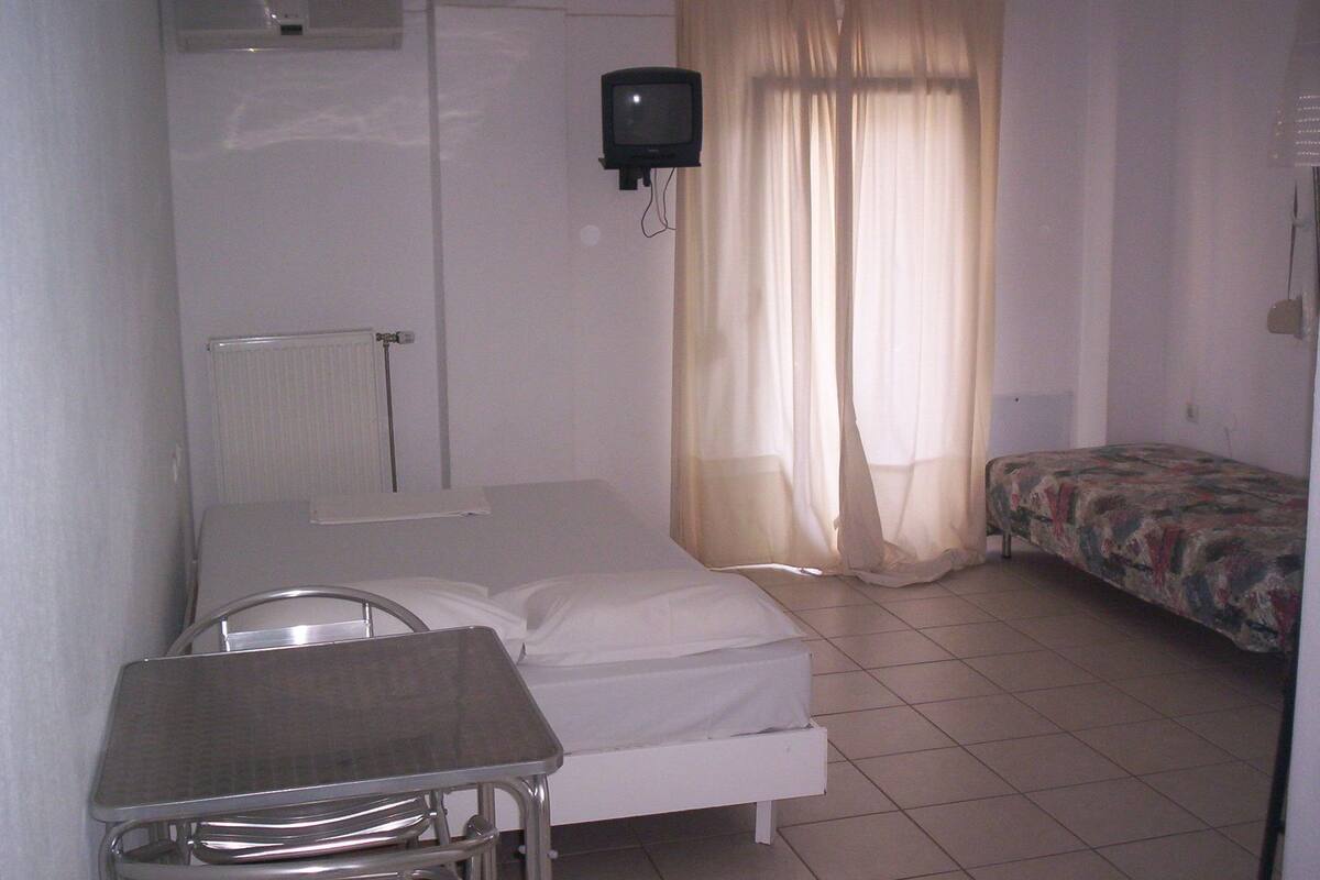 Ein schlichter, einfach eingerichteter Raum mit einem Bett, einem Tisch, einem Stuhl, einem Fernseher und einer Heizung.