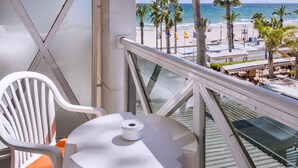Ein Balkon mit weißem Stuhl und Tisch, der auf einen Strand mit Palmen und einen klaren Blick auf den Ozean blickt.