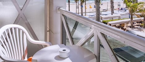 A balcony with a white chair and table, overlooking a beach with palm trees and a clear view of the ocean.
