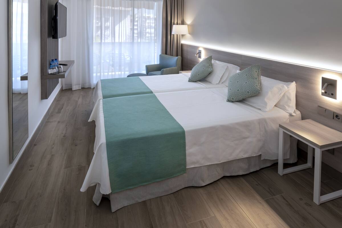 Habitación de hotel con una cama grande, una mesita de noche, una silla, un televisor en la pared y una ventana con cortinas.