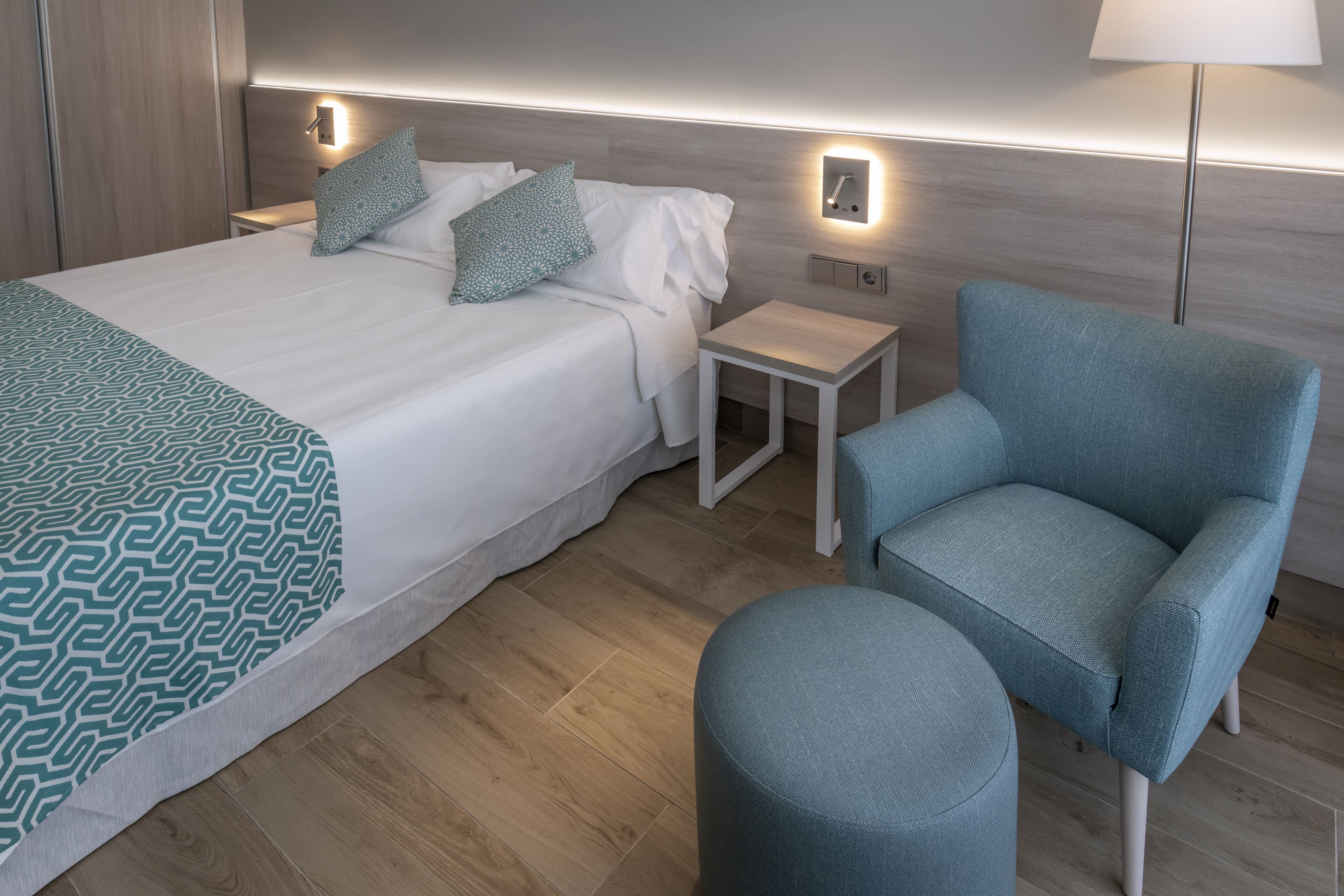 A neatly arranged hotel room with a large bed, a blue armchair, a small table, and a lamp.