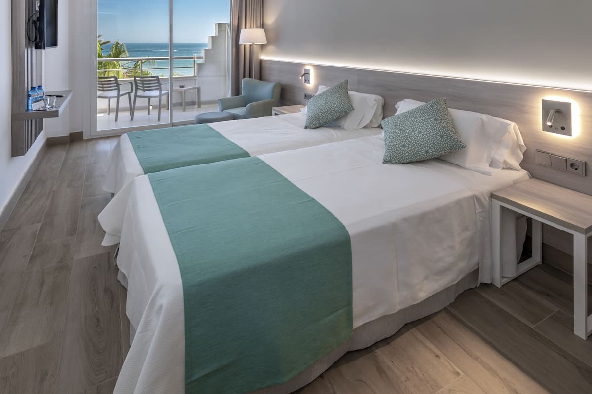 Una habitación de hotel con dos camas, un balcón con vistas al mar y una pequeña zona de estar con sillas.